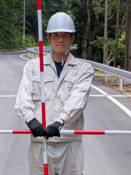 ヘルメットと作業着を着た職員が林のそばの道路で赤と白の棒を胸の高さで横に持って立っている